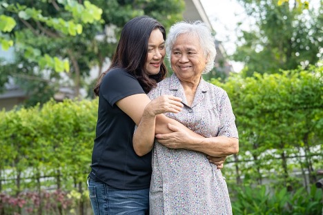 A younger woman walking arm in arm with an older woman along a tree-lined path.