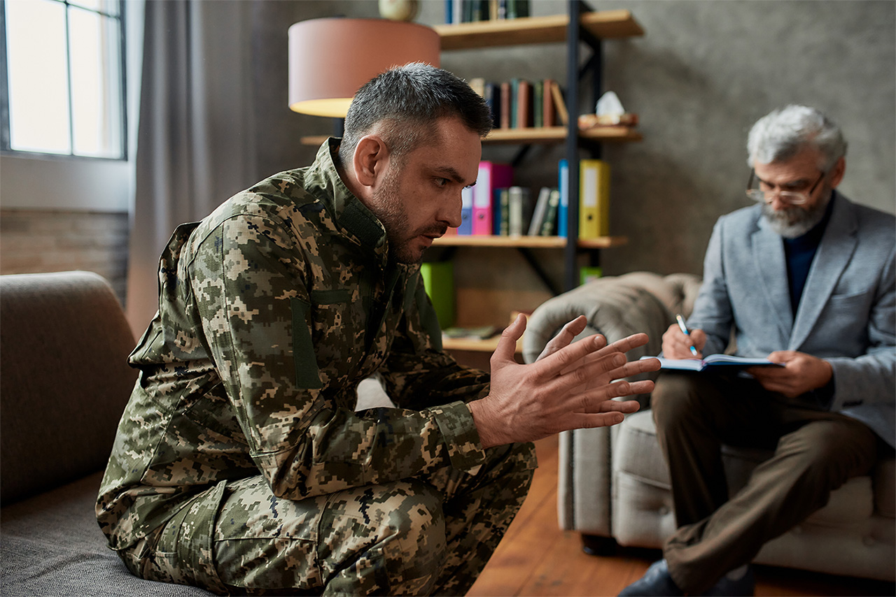 A man in military fatigues talking with a clinician who is taking notes on a clipboard.